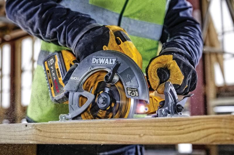 Vista de cerca de una persona usando una sierra circular inalámbrica DEWALT 60V FLEXVOLT para cortar un trozo de madera. La persona lleva guantes de trabajo y un chaleco de alta visibilidad dentro de un sitio de construcción.