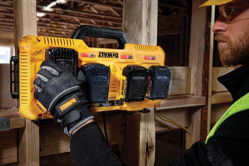 Una persona con guantes de trabajo instala o retira baterías de un cargador múltiple amarillo DEWALT montado en una viga de madera en un sitio de construcción.