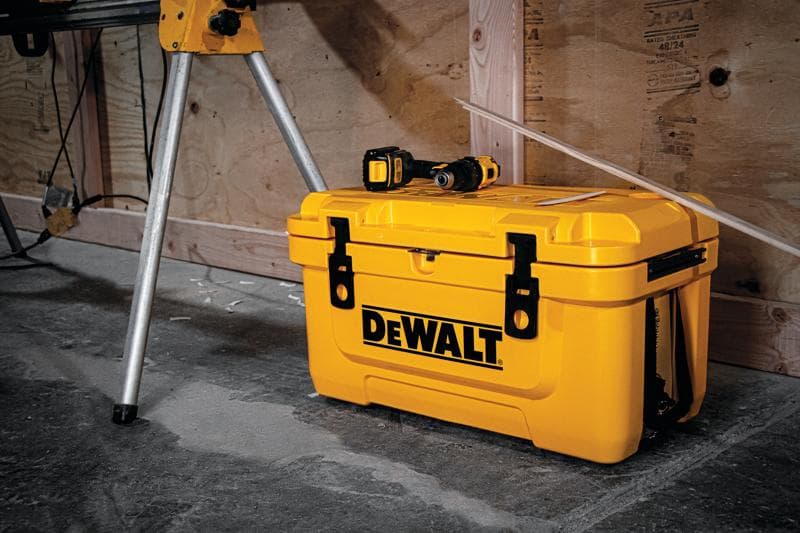 Una caja de herramientas amarilla DEWALT colocada sobre un suelo de concreto, con un taladro eléctrico encima. La caja está cerca de patas metálicas y una pared de madera en un taller.