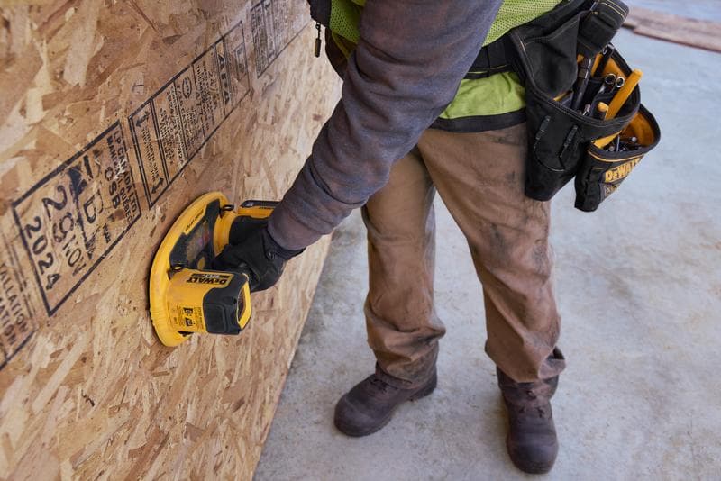 Un trabajador con pantalones marrones y guantes protectores usa una herramienta portátil DEWALT sobre una gran lámina de tablero de partículas OSB en una obra en construcción. El trabajador lleva un cinturón de herramientas con varias herramientas de mano.