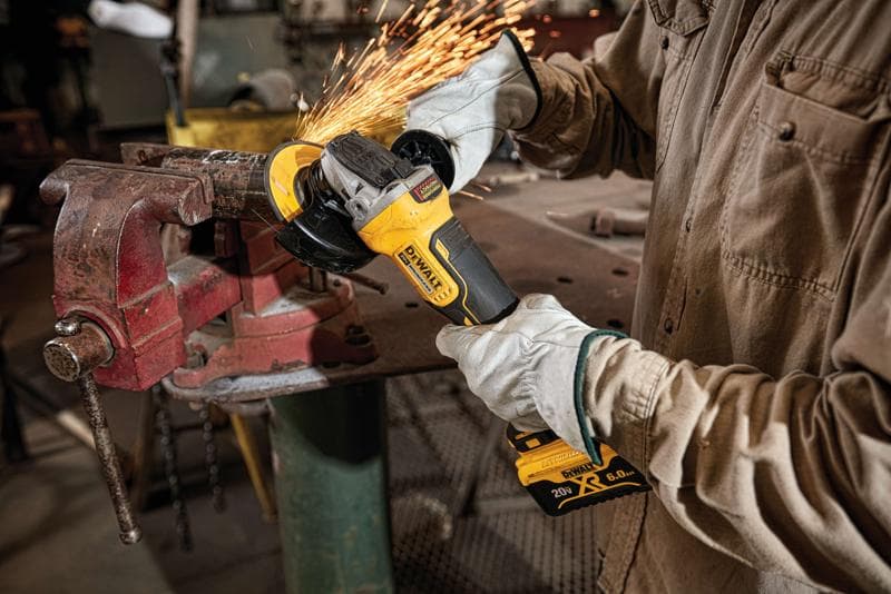 Persona usando guantes protectores utiliza una amoladora angular inalámbrica DEWALT, modelo DCG413R2, para cortar o pulir metal sujeto en un tornillo de banco, con chispas en un taller.