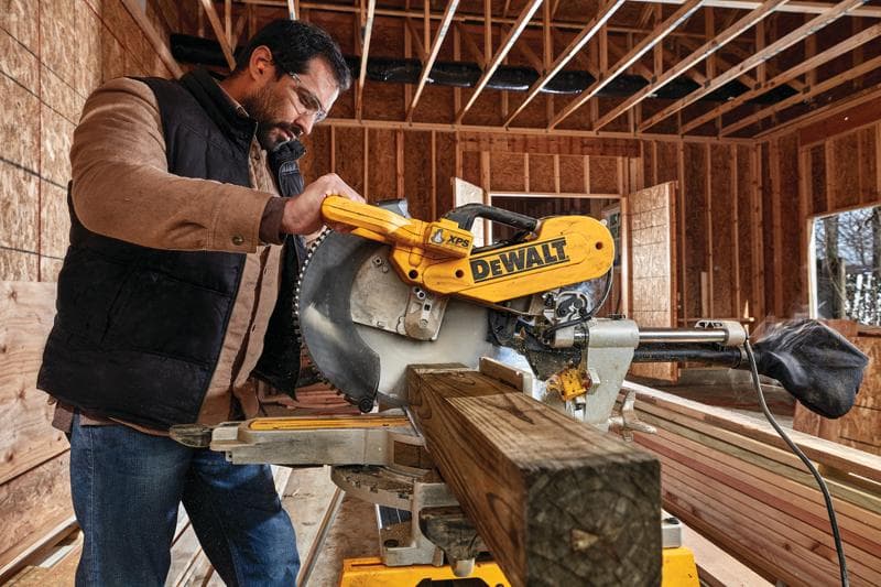 Una persona utiliza una sierra de inglete DEWALT DWS780 para cortar una viga de madera en un sitio de construcción. La sierra es amarilla y muestra el logotipo de DEWALT de manera destacada. El fondo muestra una estructura de madera expuesta y paredes sin terminar.