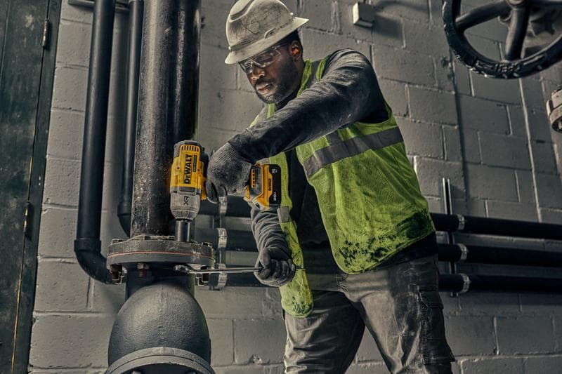Un trabajador con casco y chaleco de seguridad utiliza una herramienta eléctrica amarilla DEWALT para asegurar los pernos de una tubería industrial grande dentro de una instalación.