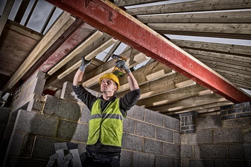 Un trabajador de la construcción con casco amarillo y chaleco reflectante utiliza un taladro inalámbrico DEWALT para fijar vigas de madera a una viga de acero roja dentro de un edificio en construcción. El rostro del trabajador está difuminado.