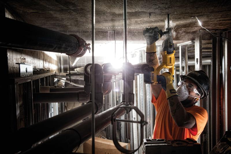 Una persona con equipo de seguridad utiliza una herramienta eléctrica amarilla DEWALT para perforar un techo de concreto en un entorno industrial con tuberías y estructuras metálicas.